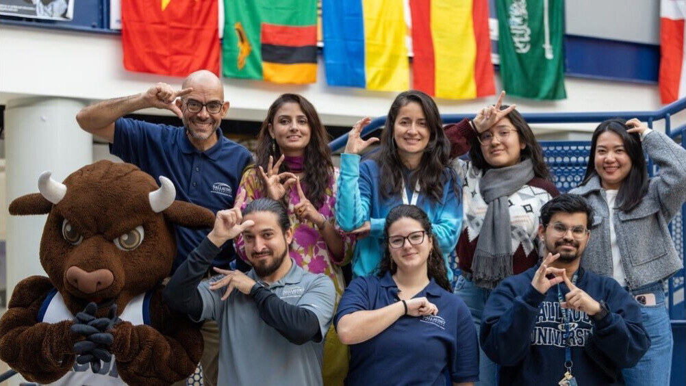 A photo of attendees at the Global Gallaudet Celebration, as well as the Gallaudet Bison Mascot. Each person is showing the sign names of international countries, such as Pakistan, Columbia, Spain, and the United States.