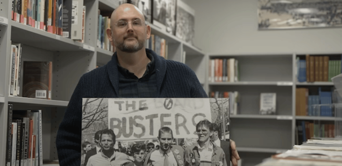 A photo of smiling Gallaudet Archivist Jim McCarthy. Jim is standing in front of library shelves. He is holding a photo of the Deaf President Now protests. The photo shows three students, with a sign in the background saying, "The Board Busters".