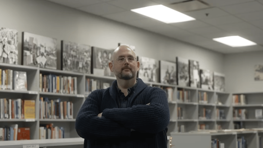 A photo of Gallaudet Archives Director Jim McCarthy in the Gallaudet library. Jim is smiling, with his arms crossed. He is bald, with salt and pepper facial hair and glasses. Behind him are shelves with books, with large photos on the top of the shelves.