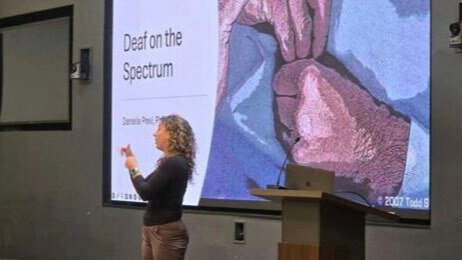A photo of Dr. Danielle Previ presenting at the National Conference on Autism in Deaf Education. Behind Dr. Previ is a projected slide with the title, “Deaf on the Spectrum”.