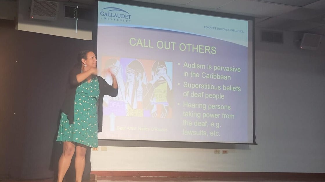 A photo of Dr. Danielle Thompson presenting on stage. Behind Dr. Thompson is a projected slide with the Gallaudet logo and text: “Call Out Others / Audism is pervasive in the Caribbean / Superstitious beliefs of deaf people / Hearing persons taking power from the deaf, e.g. lawsuits, etc.”
