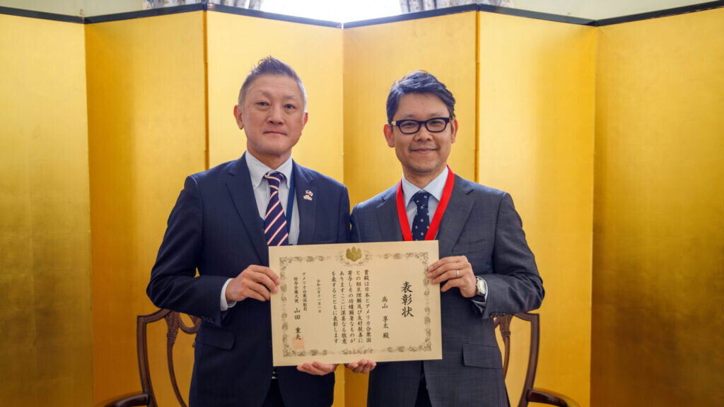A photo of Fumito Miyake, left, presenting a Japanese Ambassador’s Commendation to Dr. Kota Takayama, right. Miyake has styled grey hair and wears a dark blue suit. Dr. Takayama has parted dark hair, and is wearing glasses and a grey suit. The commendation is in Japanese kanji. The background is a bright yellow folding screen. 