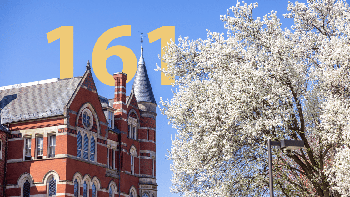 A photo of a section of College Hall on the eft, with cherry blossoms in the foreground on the right. Behind College Hall, in the blue sky, is a golden "161".