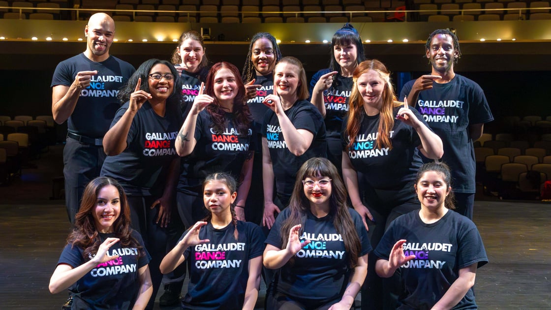 A photo of Gallaudet Dance Company members, standing in three rows of different heights. The top row is signing, "G". The middle row is signing, "D". The bottom row is signing, "C".