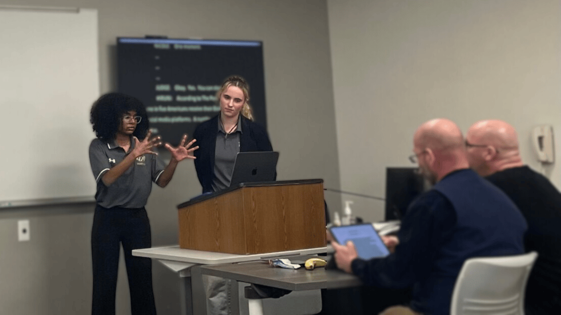 A photo of Hiruni Hewapathiranage-Mayadunne and Nicole Hicks during a debate. They are on the left side of the image, standing at a podium at the front of a classroom. Hiruni is signing on the left while Nicole watches on the right. On the right side of the image two people sit and watch..
