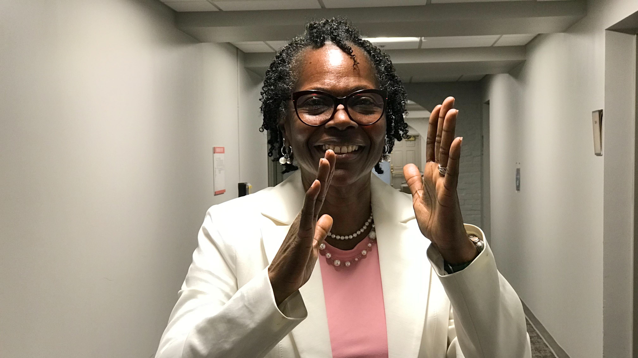 A photo of Hi5 signer Ena Harrison signing "Hi5". They have black hair. They are wearing a white blazer and pink shirt with pearls.