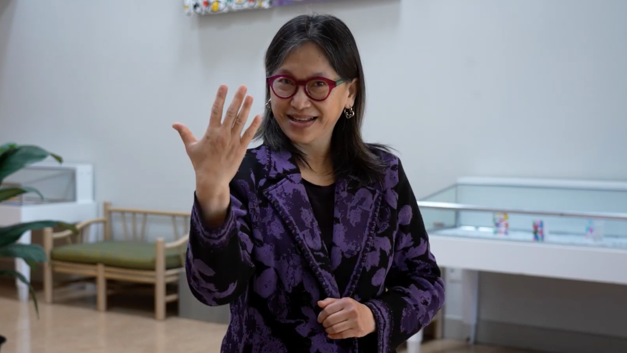A screenshot of Yiqiao Wang making a "5" handshape. Yi is wearing a jacket with a navy and black design, and glasses. They are in a lobby area.