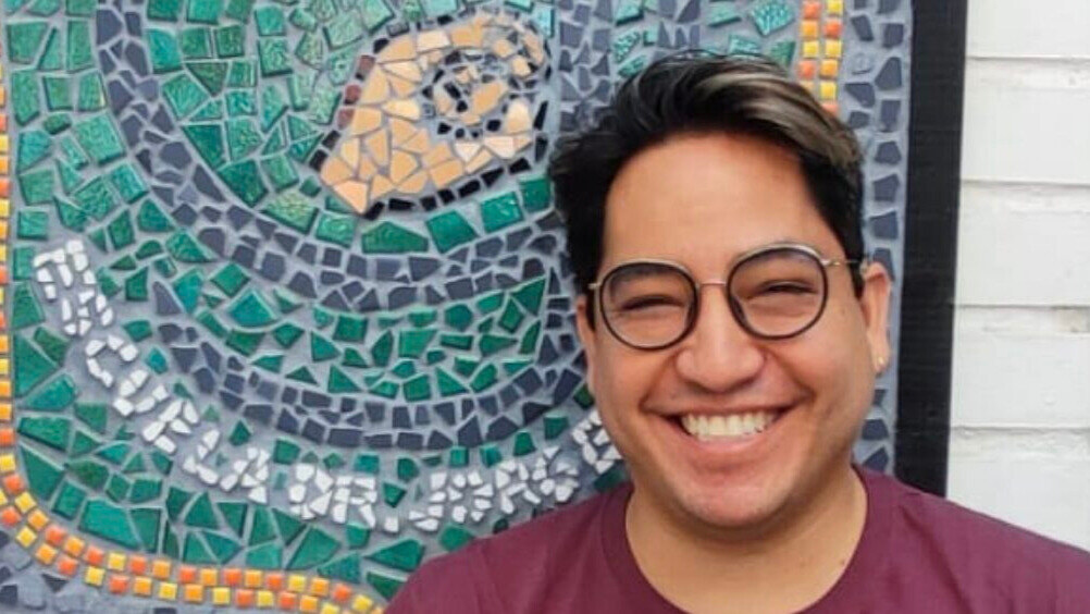 Eric Hidalgo smiles for the camera in front of a mosaic with a hand design. Hidalgo has short, dark hair with highlights and wears glasses.