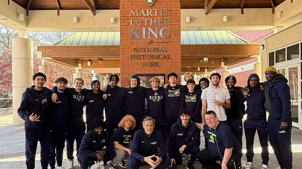 A photo of the Gallaudet Men’s Basketball team, players and staff. They are posting in front of a sign for the Martin Luther King, Jr. National Historical Park, under a large portico and in front of a building entrance.