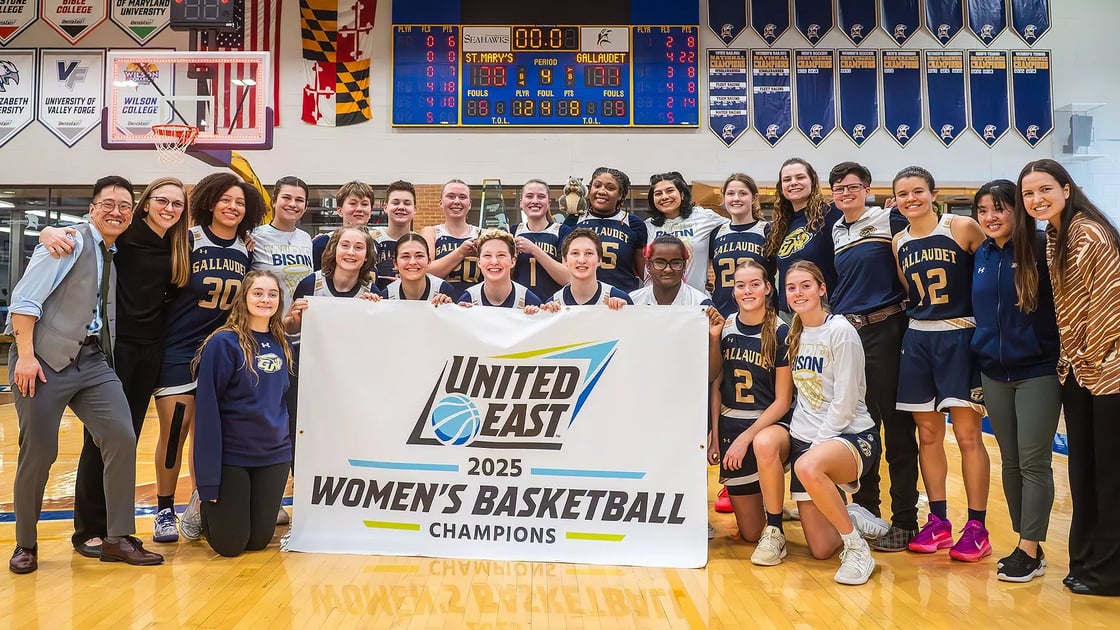 The Gallaudet Women’s Basketball players and coaches pose on a court, holding a banner that reads, “United East 2025 Women’s Basketball Champions”.