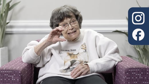 A photo of an elderly person sitting in a chair, signing the letter "C" in ASL. They have short, dark gray hair. They wear a white sweatshirt. The background is a plain wall with green plants on either side of the screen. On the top right of the image are Instagram and Facebook logos.
