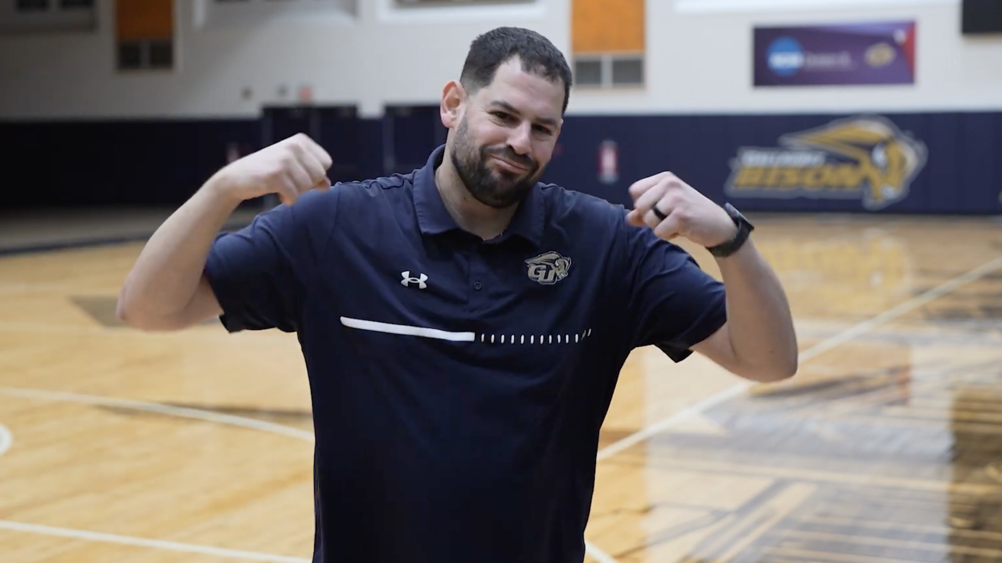 A screenshot of Bruce Saperstein, signing "Bison" in the Gallaudet Field House, with a basketball court in the background.