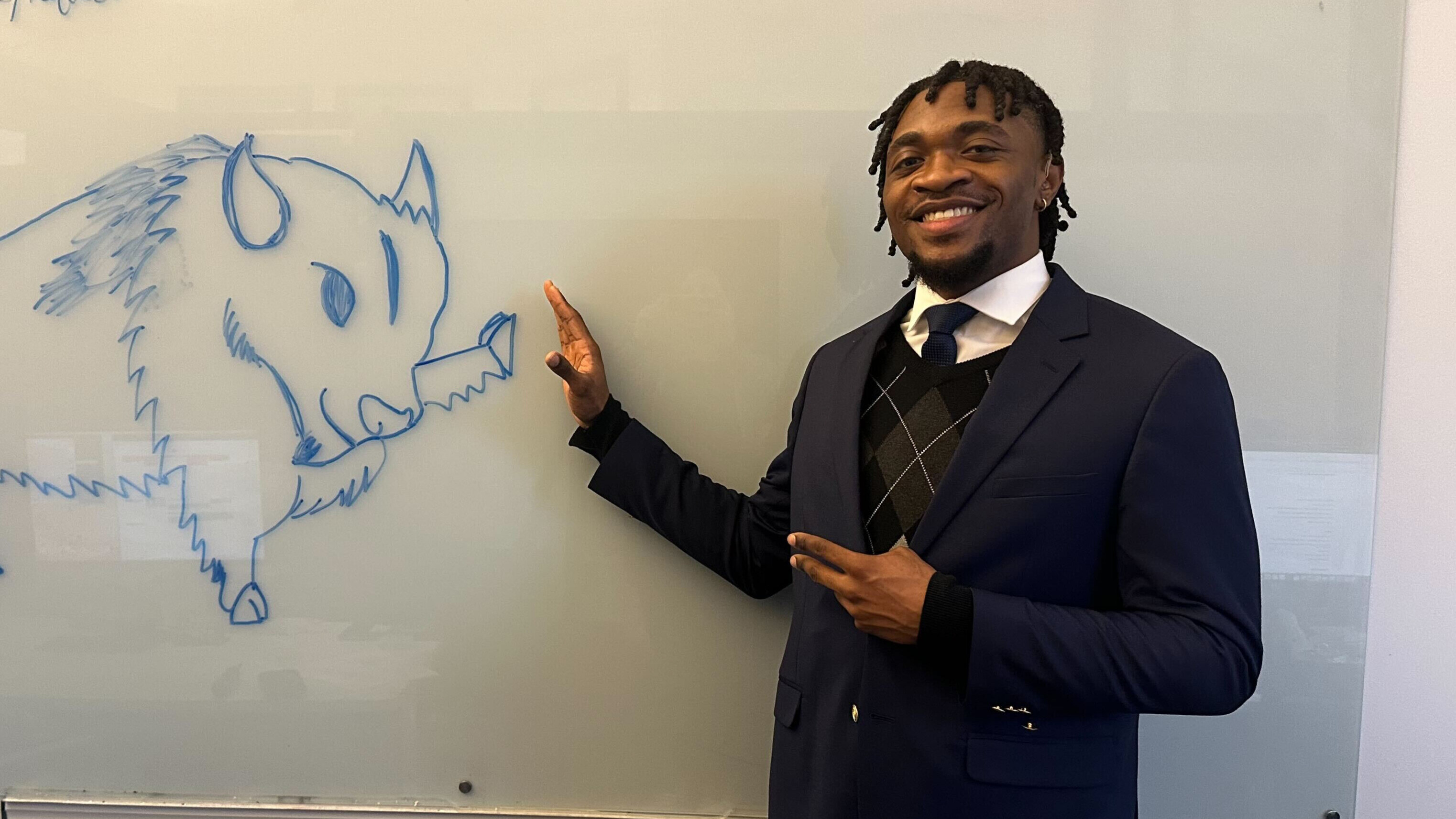 A photo of Enow Otto pretending to high five a drawing of a Bison on the whiteboard behind him. On the right side, Enow is smiling, wearing a blue blazer, white shirt, tie, and pullover. The Bison on the left is drawn with blue ink, a sly expression on its face.