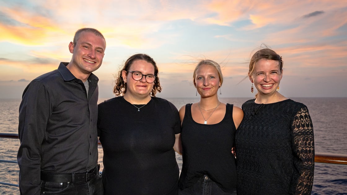 A photo of four people smiling for the camera on the deck of a ship. Second from the left is Gallaudet student Julia Bronneberg. Fellow student Macy DeGraw is to Julia’s right. They are bookended by ASL interpreters on the left and right. In the background is the ocean, with a sunset-adjacent sky.