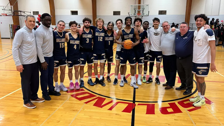 A photo of the Gallaudet men’s basketball players and coaches standing in a row on a basketball court. In the middle, Blessed Mbogo smiles and holds a basketball.