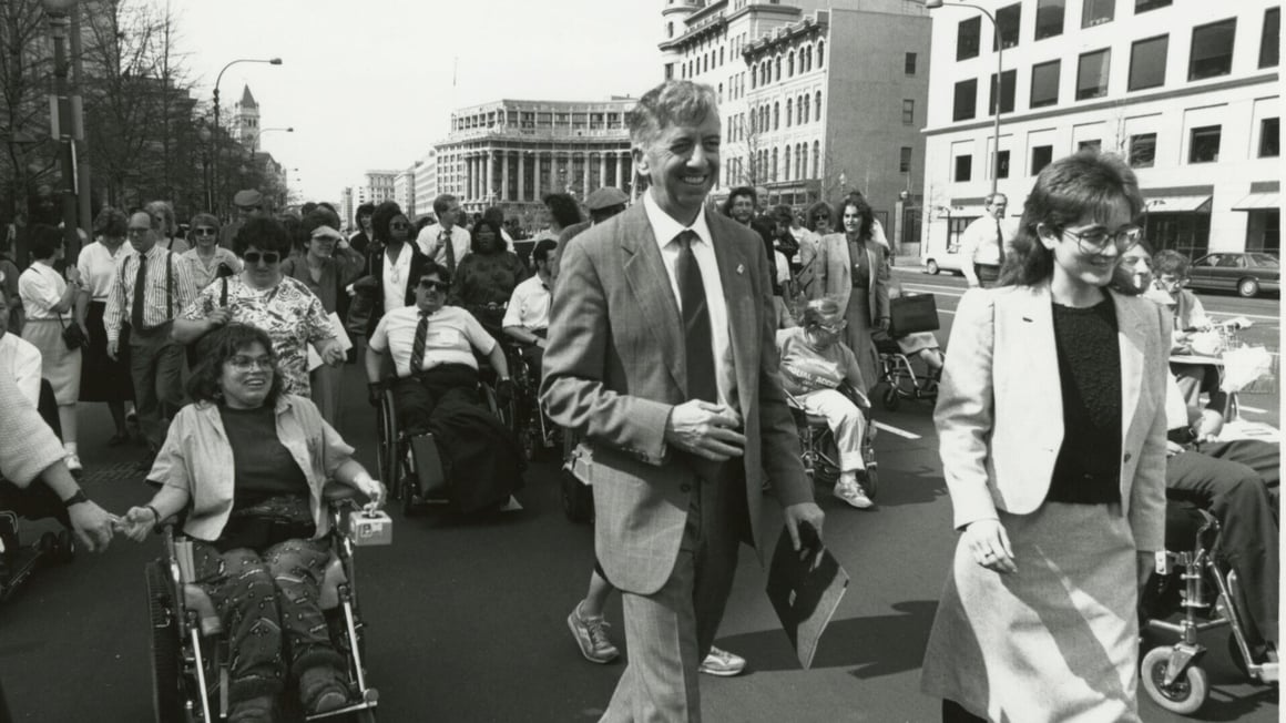 A black and white photo of disability rights activists marching on the street in support of the Americans with Disabilities Act (ADA), before it was passed. In the front, on the left, is Judy Heumann in an electric wheelchair. To Heumann’s left is former Gallaudet President I. King Jordan.
