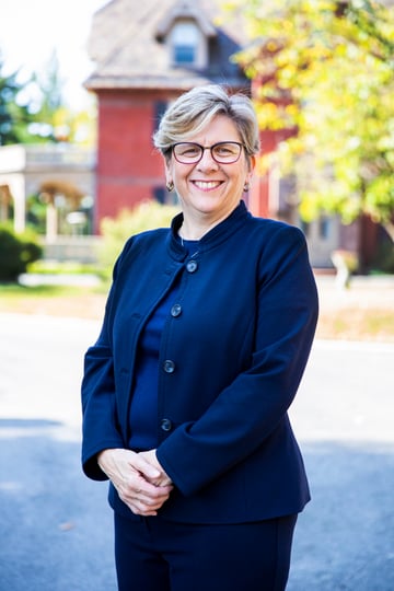 President Cordano, smiling and wearing a black business suit, stands in front of House One. Behind her is a leafy tree, brick house with a wraparound patio, driveway and a lawn.