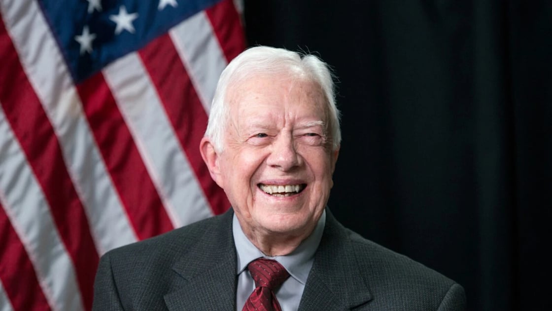 A photo of former US President Jimmy Carter, smiling. He has thin white hair and wears a grey suit. There is an American flag in the left half of the background.