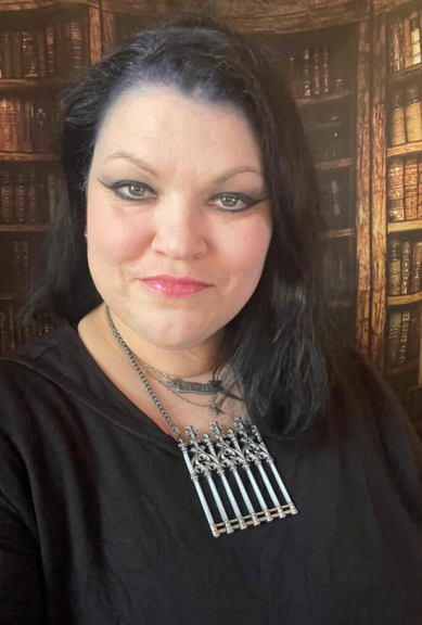 Sharon Pajka, smiling slightly and wearing a black shirt and an ornate silver necklace, is standing in front of a bookshelf.
