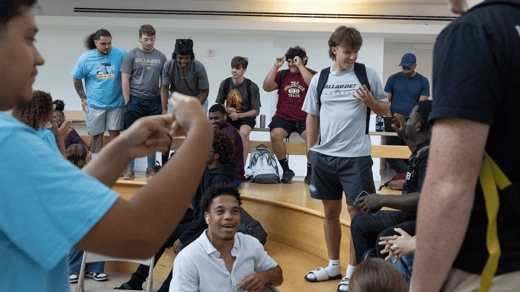 A photo of many college students hanging out in a building atrium. In the right center, a student in a t-shirt and with a backpack stands on wooden seating, smiling at a peer. In the bottom center, a seated student smiles at somebody offscreen. In the back, a line of students look at each other.
