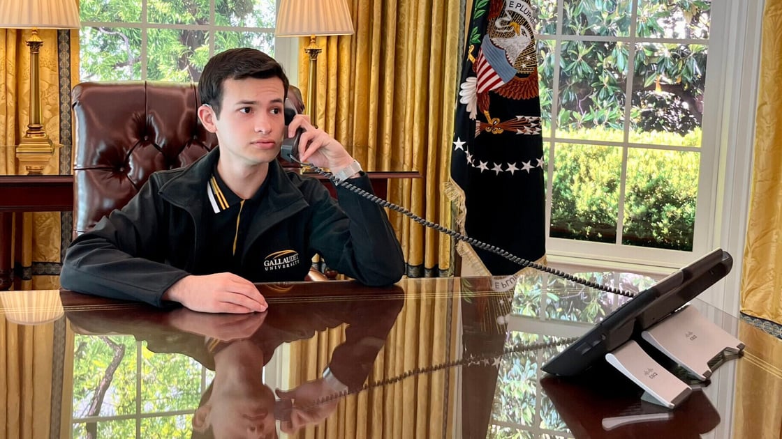 A photo of José Rodrigo Uresti sitting at the Resolute desk in the Oval Office. José has short brushed hair and a Gallaudet jacket. He has a phone in his hand and looks to the right offscreen. The desk reflects José, as well as the gold curtains and foliage visible through the windows behind José.