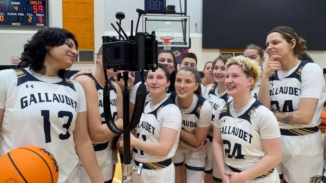 A photo of the Gallaudet women’s basketball team huddled around a camera and smiling. The players are wearing their white jerseys.