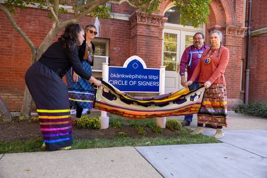 Four people wearing colorful attire unveil the new building sign for Building 103. They are holding a flag and lowering it to show the new name. 
