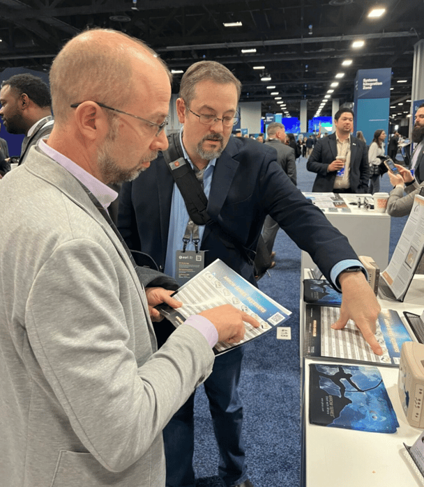 Eric Kaika is wearing a gray jacket on the left and Erick Posner, attired in blue jacket/jeans points to a paper on a booth. Both are bespectacled.  