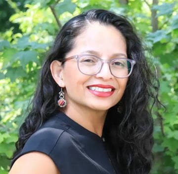 Norma Morán, wearing glasses and a black shirt, stands in front of green foliage.